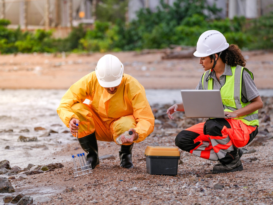 Two environmental engineers in safety gear work at a shoreline near gas storage tanks. One collects water samples, while the other records data on a laptop during site inspection.