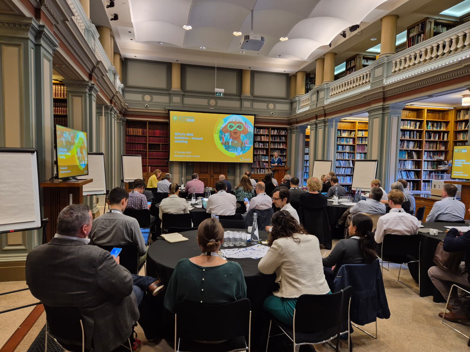 The audience sits at tables watching a screen in the Burlington House library with the Sustainable PLFs report cover on the screen
