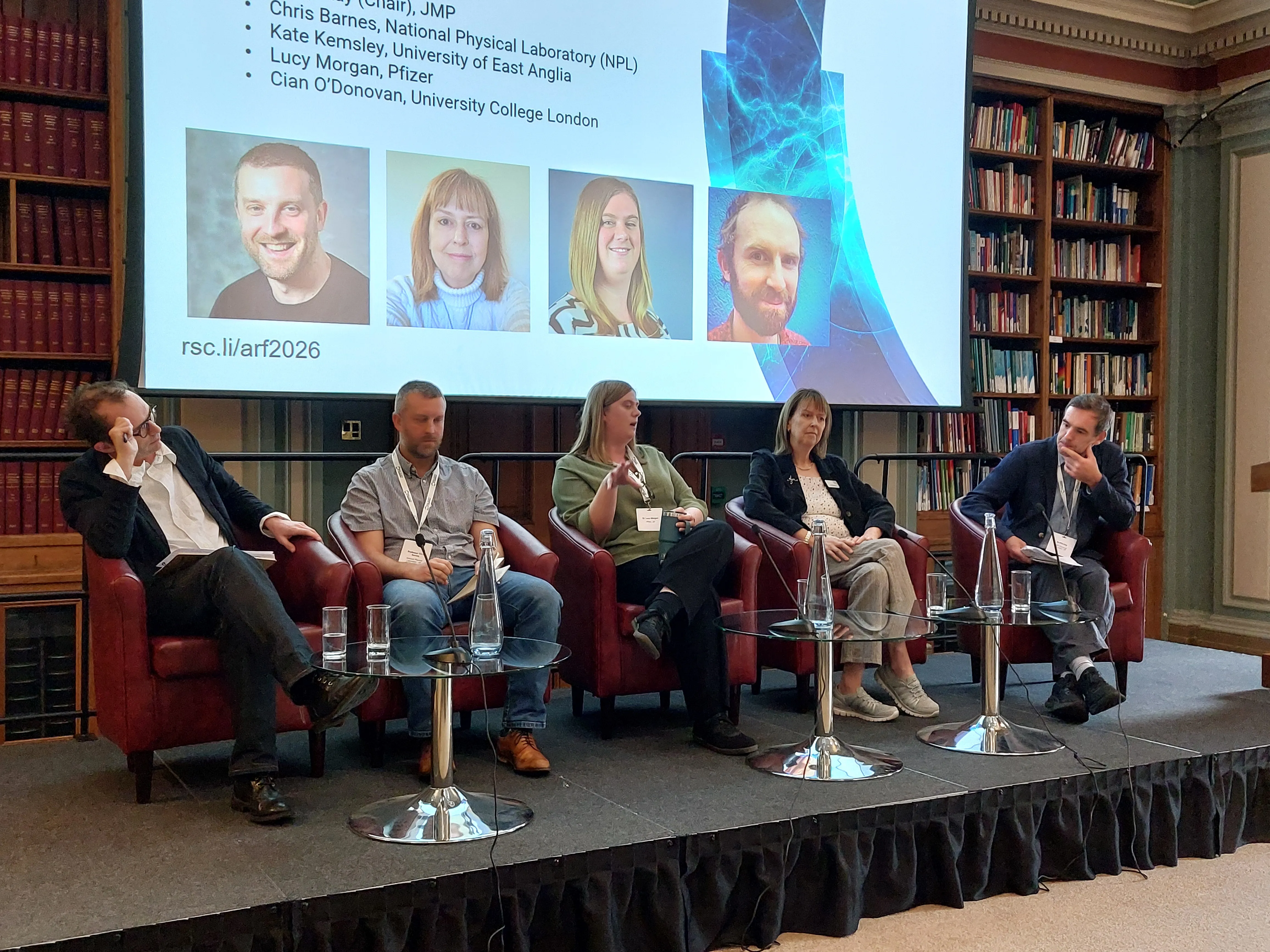 Analytical Research Forum - panel session Five people sit on a stage in front of a screen with their faces on it
