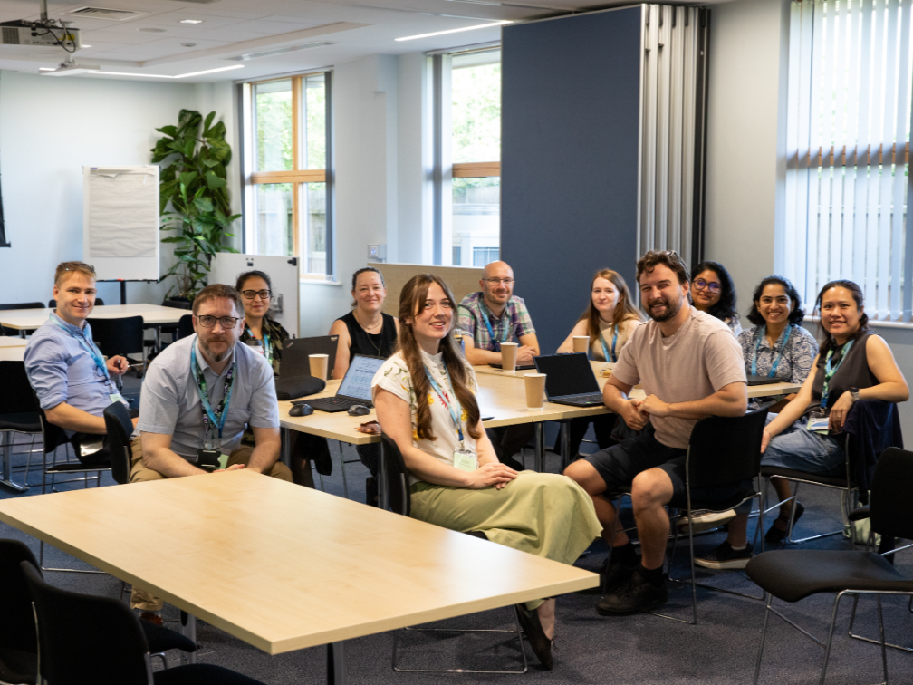 A group of approximately ten diverse colleagues are smiling and posing together around office desks in a bright, modern conference room or classroom setting.