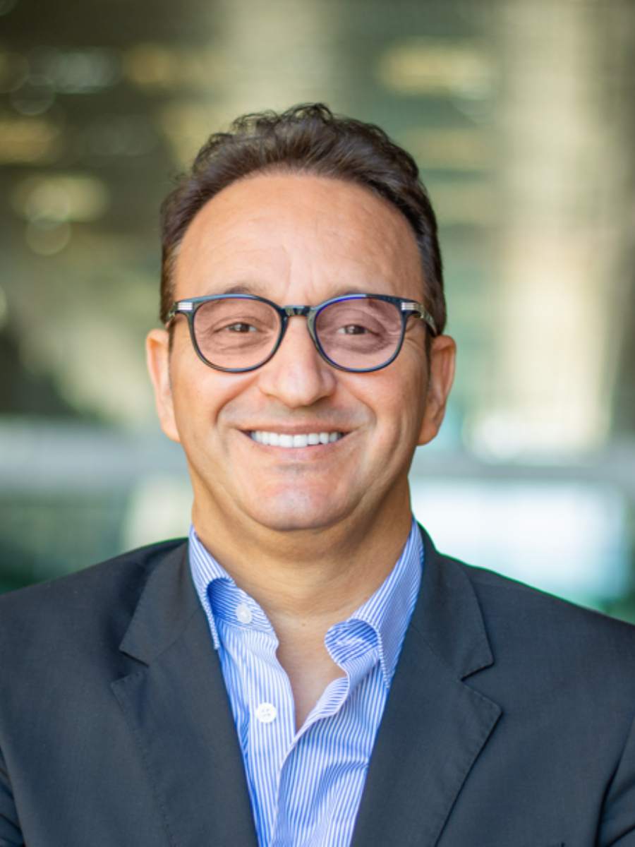 Mohamed Eddaoudi smiling widely to camera wearing glasses and suit jacket and blue shirt